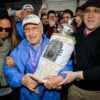 Elderly man in a blue jacket and cap holding an ornate Torah scroll as a cheering crowd surrounds him at a celebration.
