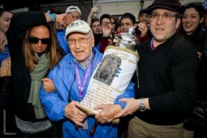 Elderly man in a blue jacket and cap holding an ornate Torah scroll as a cheering crowd surrounds him at a celebration.