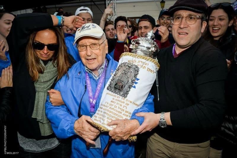 Elderly man in a blue jacket and cap holding an ornate Torah scroll as a cheering crowd surrounds him at a celebration.