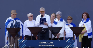 Man speaks into a microphone at a podium, flanked by six adults on stage, all wearing white tops and blue scarves; banner shows 'DANIEL LOUZ' and 'HOLOCAUST SURVIVOR'.