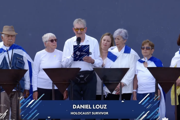 Man speaks into a microphone at a podium, flanked by six adults on stage, all wearing white tops and blue scarves; banner shows 'DANIEL LOUZ' and 'HOLOCAUST SURVIVOR'.