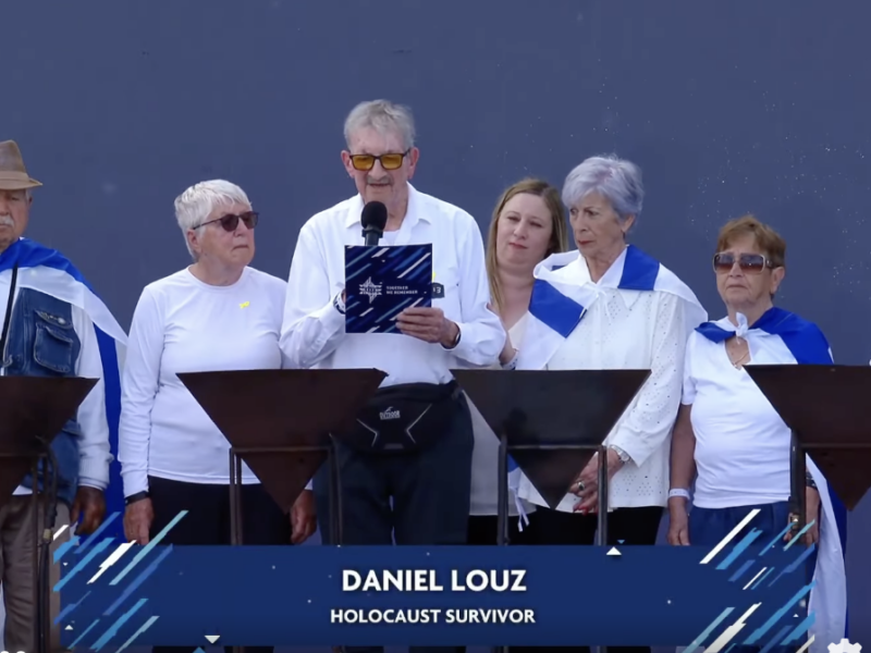 Man speaks into a microphone at a podium, flanked by six adults on stage, all wearing white tops and blue scarves; banner shows 'DANIEL LOUZ' and 'HOLOCAUST SURVIVOR'.