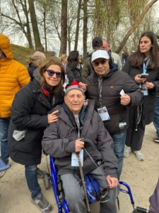Elderly man in a wheelchair flanked by a smiling woman at an outdoor event with trees in the background.