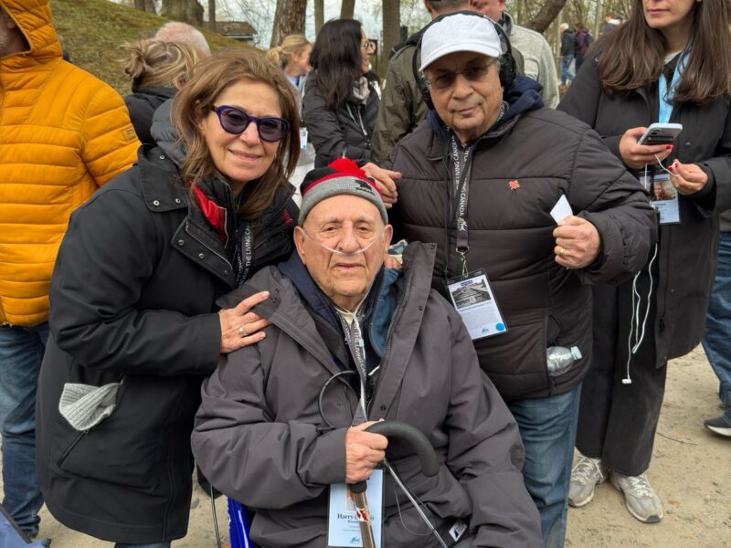 Elderly man in a wheelchair flanked by a smiling woman at an outdoor event with trees in the background.