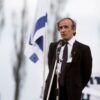 Man in a dark suit speaks into a microphone at an outdoor event, with a blue-and-white flag in the background.