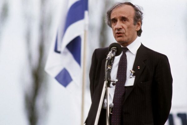 Man in a dark suit speaks into a microphone at an outdoor event, with a blue-and-white flag in the background.