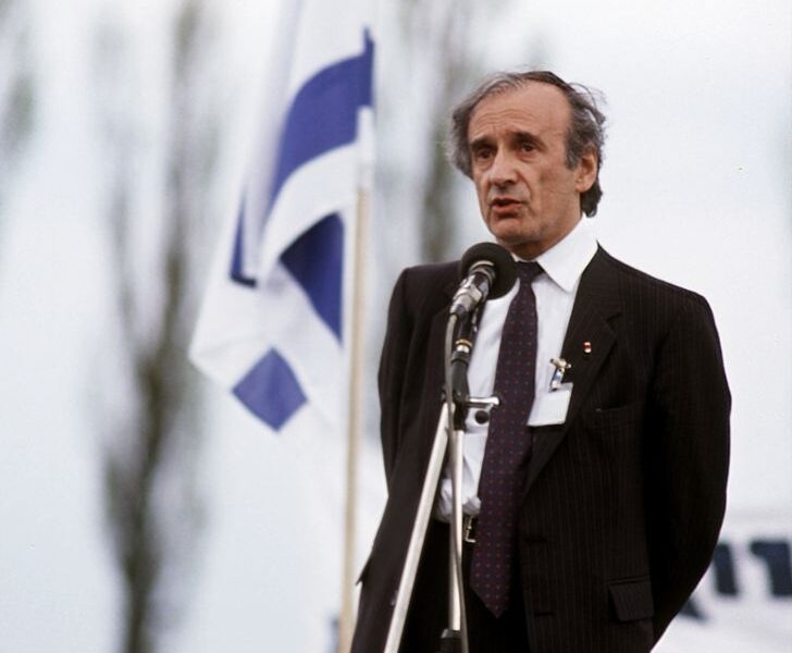 Man in a dark suit speaks into a microphone at an outdoor event, with a blue-and-white flag in the background.
