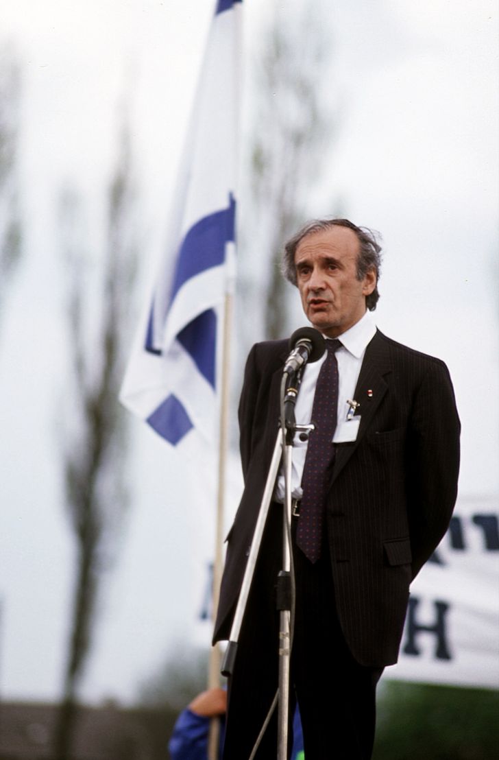Man in a dark suit speaks into a microphone at an outdoor event, with a blue-and-white flag in the background.