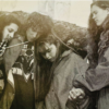 Four young musicians form a tight circle, heads bowed, during a performance with a microphone stand nearby.