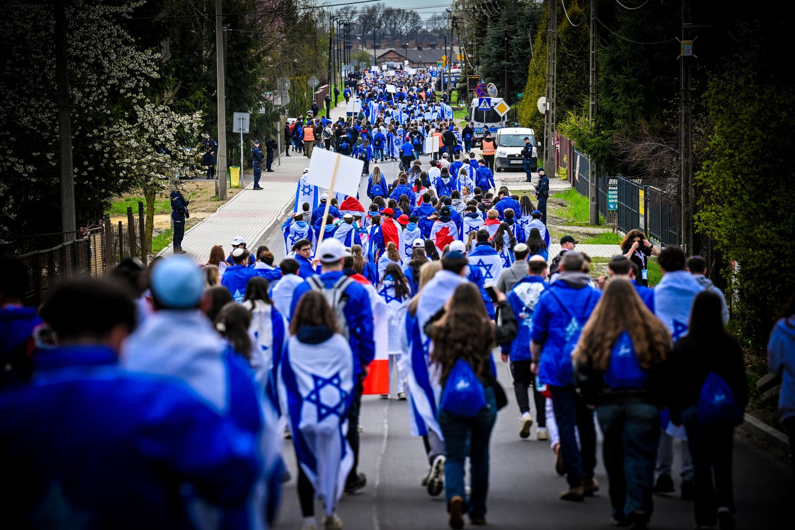 A long procession of March of the Living participants in blue jackets and flags on the road from Auschwitz to Birkenau