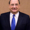 Smiling man in a dark suit and blue tie posing against a beige wall