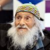 Older man with a long white beard wearing a colorful knit cap and dark jacket, looking off to the left.