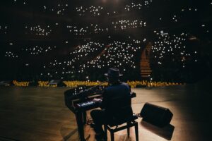 Pianist wearing a hat playing a grand piano on a dimly lit stage, with small bright audience lights in the background.