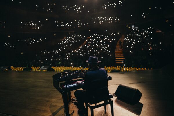 Pianist wearing a hat playing a grand piano on a dimly lit stage, with small bright audience lights in the background.