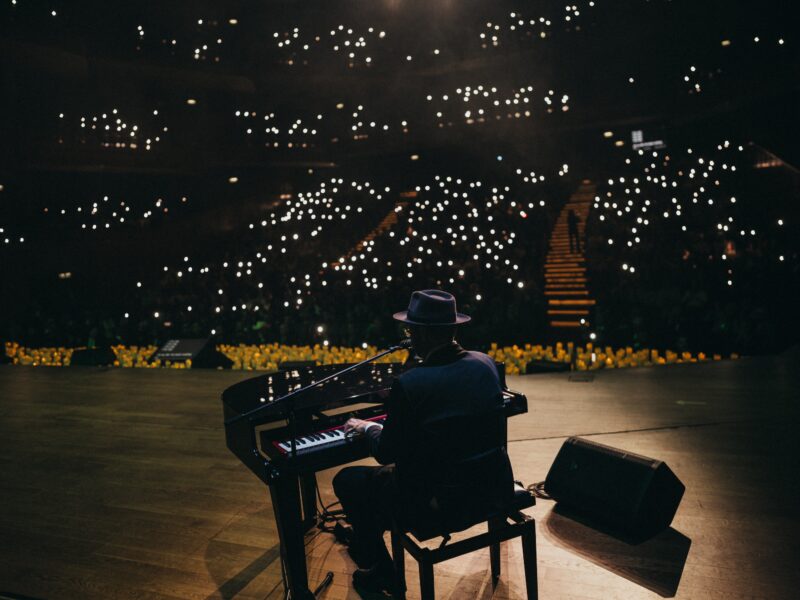 Pianist wearing a hat playing a grand piano on a dimly lit stage, with small bright audience lights in the background.
