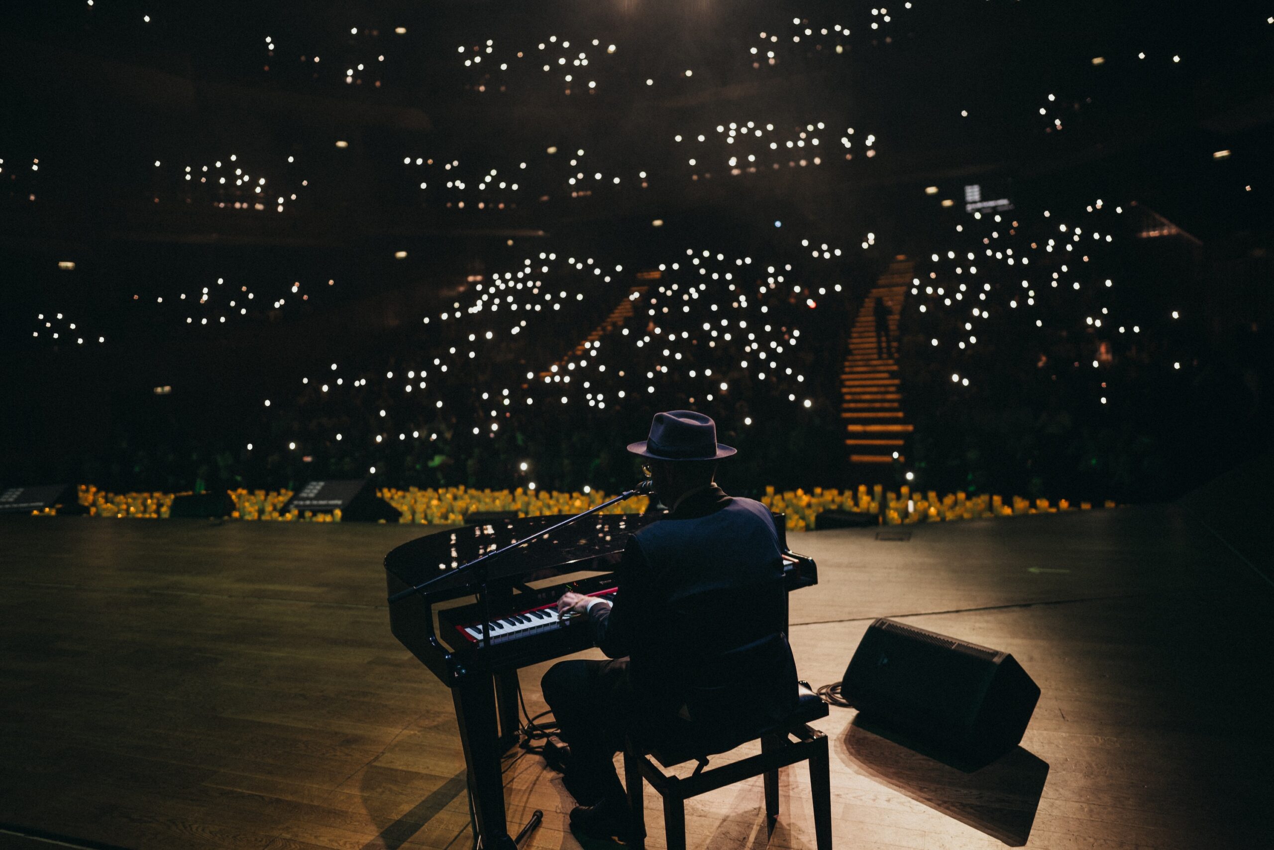 Pianist wearing a hat playing a grand piano on a dimly lit stage, with small bright audience lights in the background.